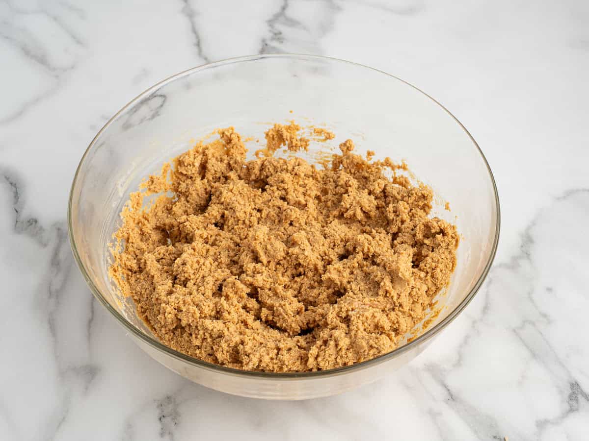A clear glass bowl filled with light brown cookie dough—perfect for a no bake peanut butter bars recipe—sits on a white marble countertop.