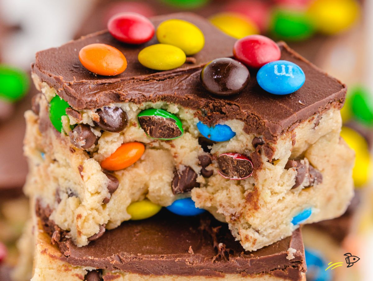 Close-up of thick cookie dough bars topped with a smooth chocolate ganache and colorful mini M&M’s, showing the chewy cookie dough center packed with chocolate chips and candy pieces.