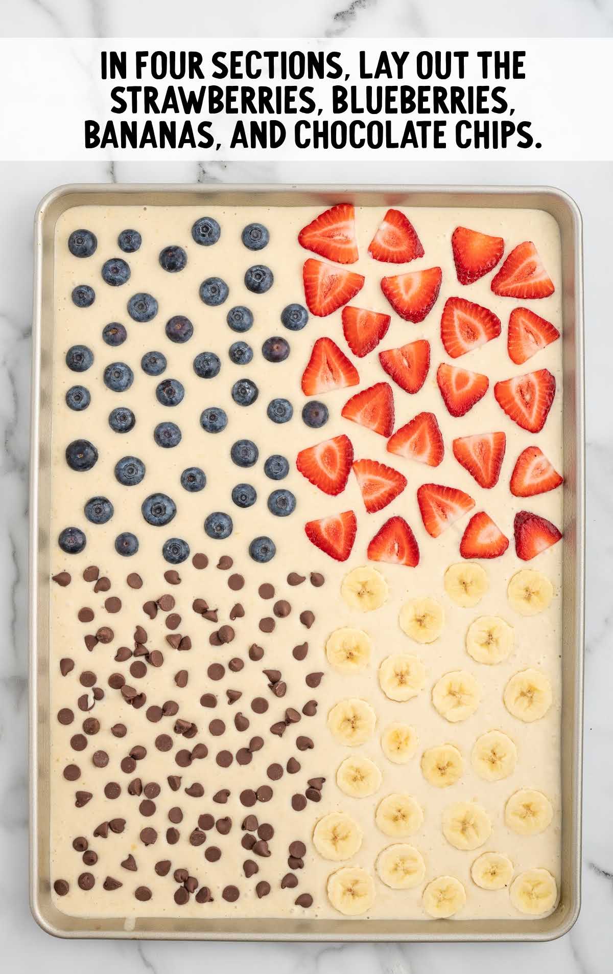 pancake batter topped with strawberries, blueberries, sliced bananas, and chocolate chips in a sheet pan