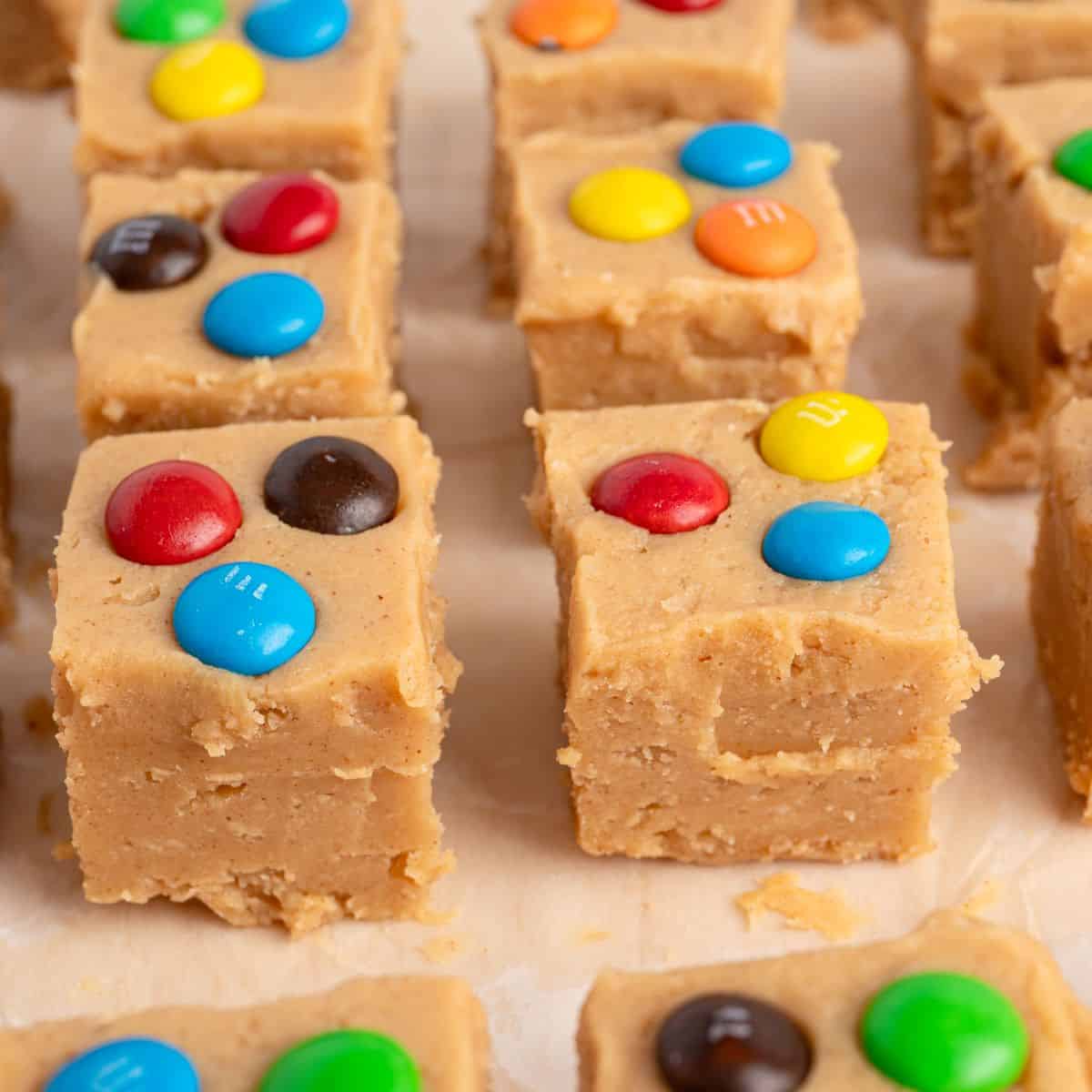 Squares of Peanut Butter Fudge (Microwave) are topped with colorful candy-coated chocolate pieces and arranged on parchment paper. The fudge has a smooth, slightly crumbly texture, making this easy treat both eye-catching and delicious.