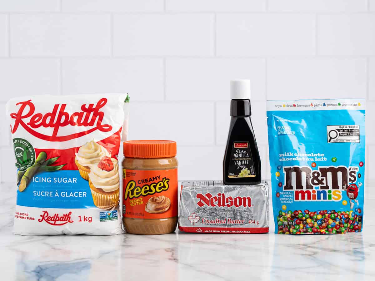 A bag of Redpath icing sugar, Reeses creamy peanut butter, Neilson unsalted butter, vanilla extract, and M&Ms minis on a white marble surfaceโperfect ingredients for an easy Peanut Butter Fudge (Microwave) Recipe.