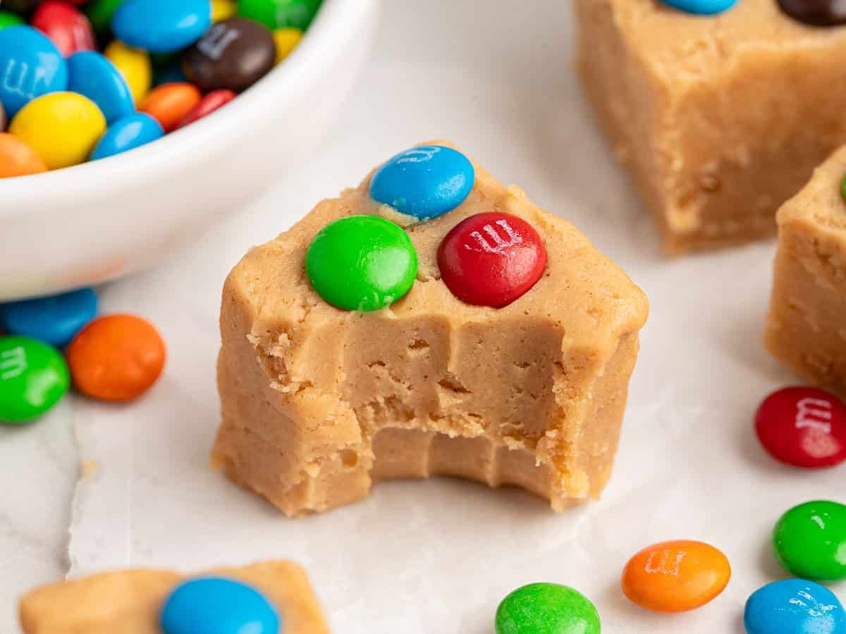 A piece of Peanut Butter Fudge (Microwave) topped with colorful candy-coated chocolates, with a bite taken out, sits on parchment paper near a bowl of more candies and other fudge pieces.