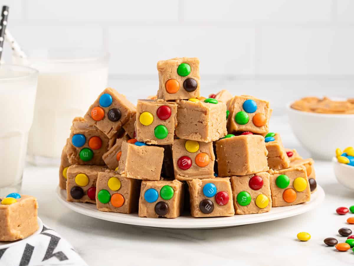 A plate stacked with square pieces of Peanut Butter Fudge (Microwave) topped with colorful candy-coated chocolates, with two glasses of milk and a bowl of peanut butter in the background.