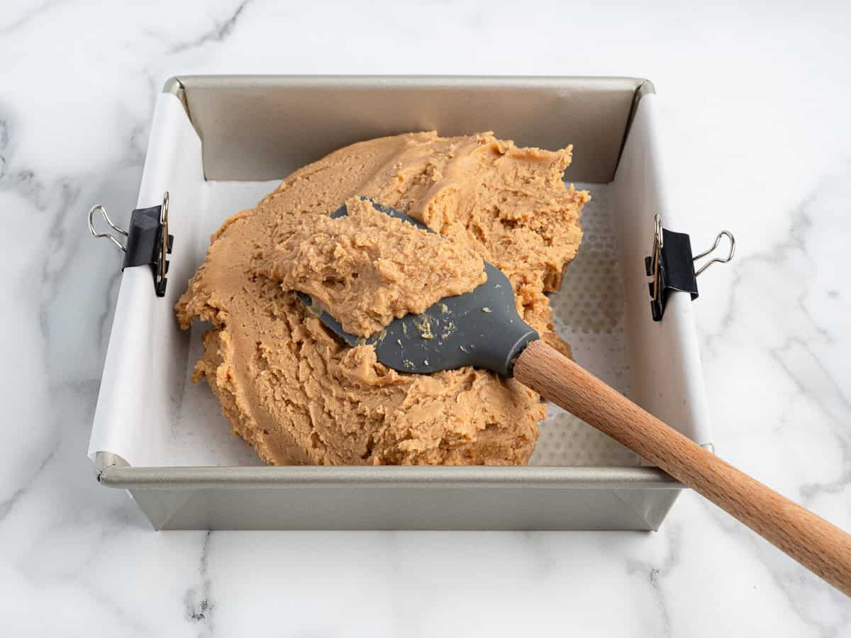 A square baking pan lined with parchment paper and held by binder clips contains thick, tan Peanut Butter Fudge (Microwave) being spread with a black spatula on a white marble surface.