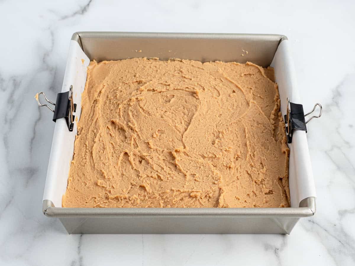 A square baking pan lined with parchment and binder clips holds an even layer of light brown Peanut Butter Fudge (Microwave) batter, resting on a marble surface.