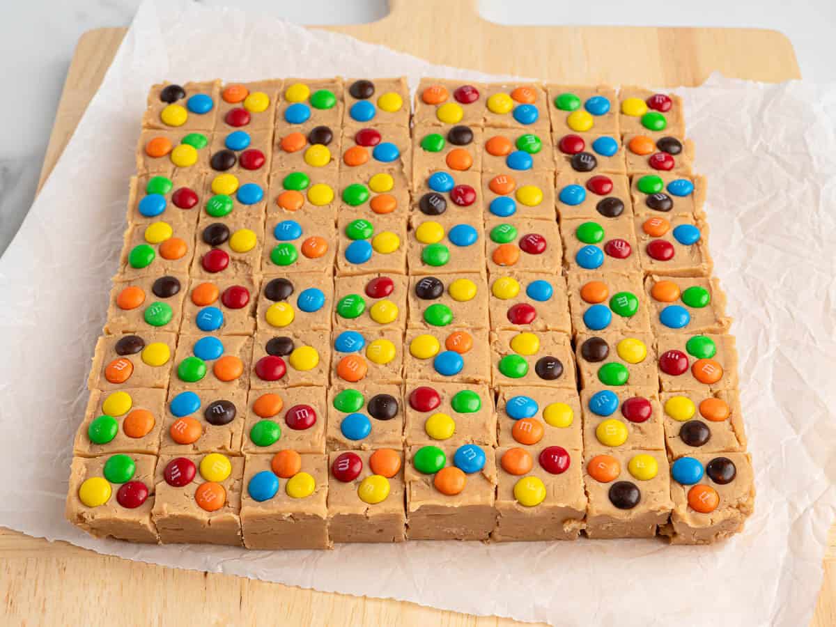 A large square block of Peanut Butter Fudge (Microwave), topped with colorful candy-coated chocolates, is cut into small squares and placed on parchment paper on a wooden surface.