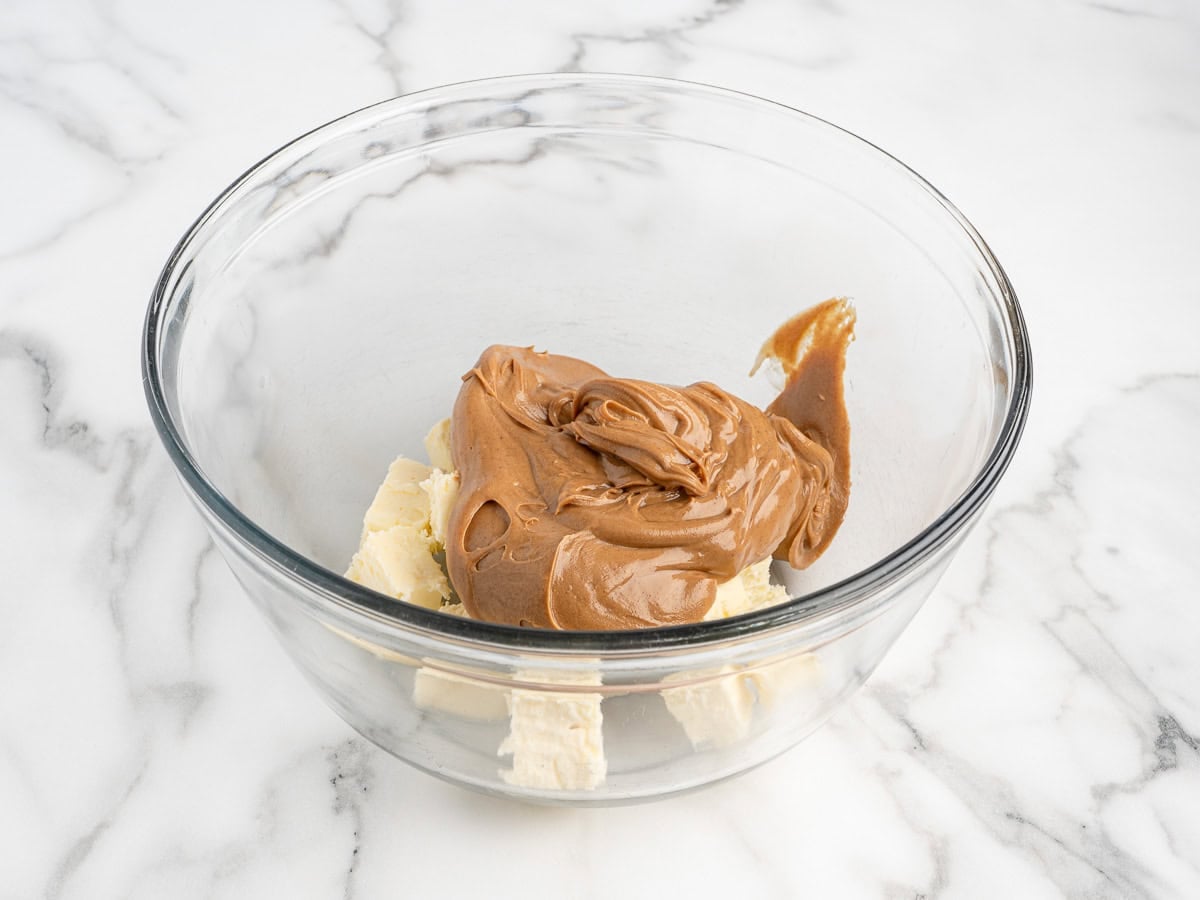 A clear glass bowl on a marble surface holds a pile of peanut butter atop chunks of white cream cheese, perfect for starting your Peanut Butter Fudge (Microwave) Recipe.