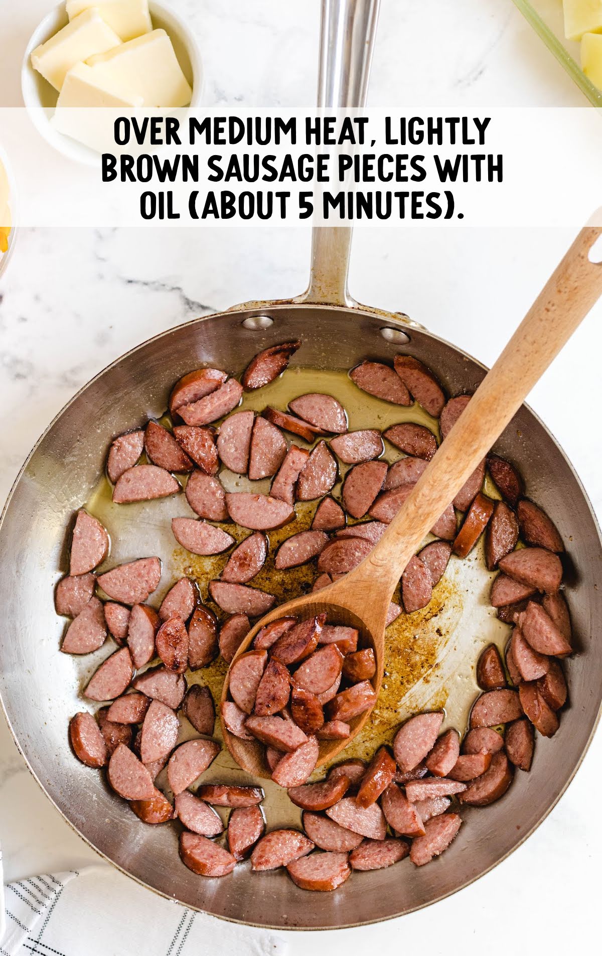 Sliced kielbasa sausage browning in a stainless steel skillet with olive oil, stirred with a wooden spoon until lightly caramelized.