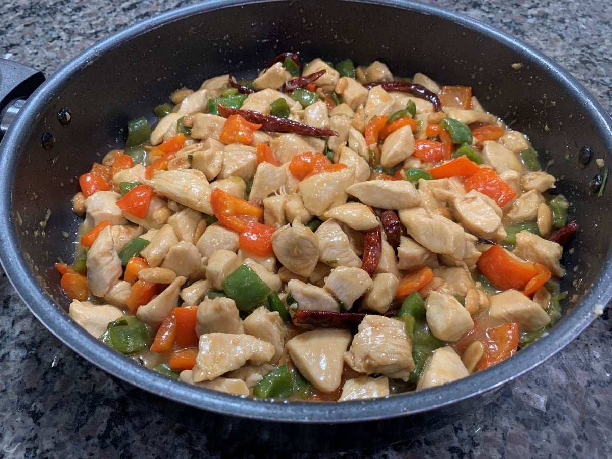 A skillet filled with homemade kung pao chicken—tender stir-fried chicken pieces, bell peppers, carrots, and dried red chilies in a savory sauce—sits atop a granite countertop.