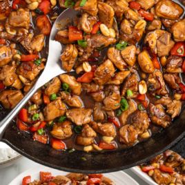 A skillet filled with homemade kung pao chicken, featuring diced chicken, red bell peppers, peanuts, and green onions in a glossy sauce. A spoon and a serving on a white plate nearby. Text reads Kung Pao Chicken, Better Than Takeout!.