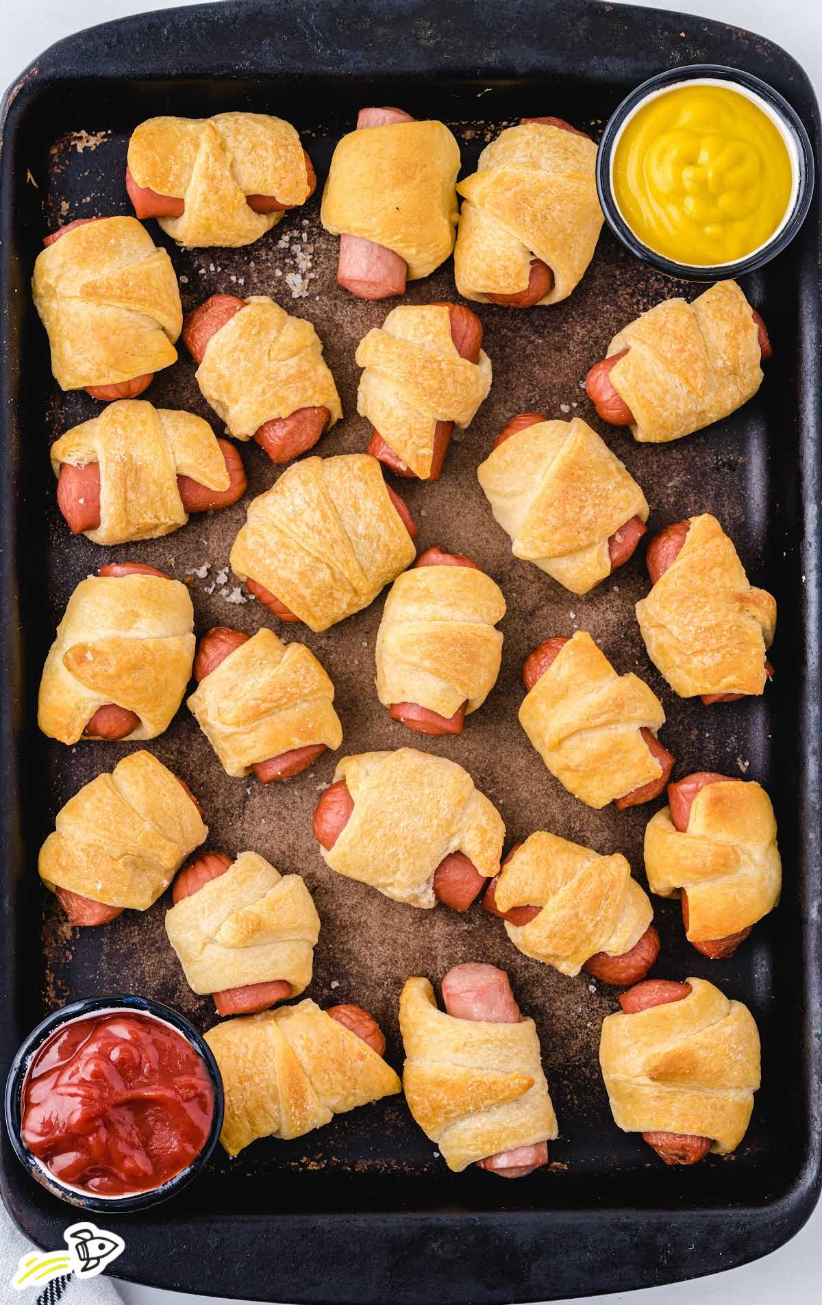 overhead shot of multiple Pigs in a Blanket on a baking dish with ketchup on the side