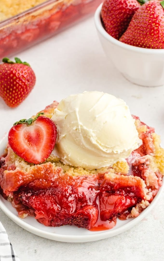 close up shot of strawberry cobbler on a white plate topped with a strawberry and vanilla ice cream