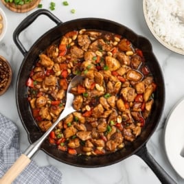 A cast iron skillet filled with the best kung pao chicken recipe—diced chicken, red bell peppers, peanuts, and glossy brown sauce—garnished with green onions. A serving spoon, rice, and side dishes are nearby.