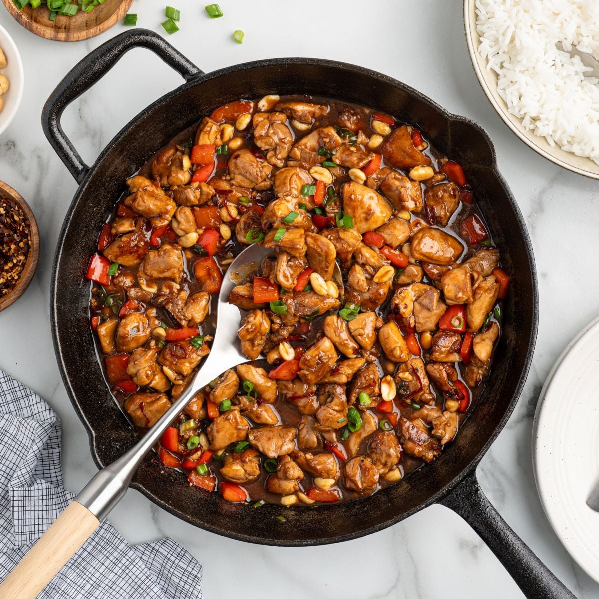 A cast iron skillet filled with the best kung pao chicken recipeโdiced chicken, red bell peppers, peanuts, and glossy brown sauceโgarnished with green onions. A serving spoon, rice, and side dishes are nearby.