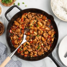 A skillet filled with kung pao chicken, garnished with green onions and peanuts, surrounded by bowls of rice, chili flakes, and silverware on a marble surface—perfect for enjoying the best kung pao chicken recipe at home.