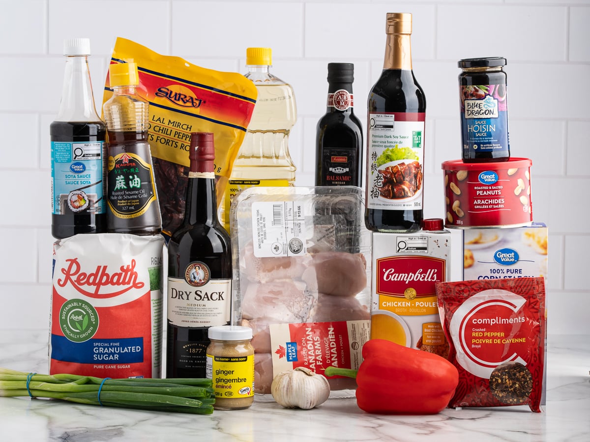 A variety of cooking ingredients for a kung pao chicken recipe, including sauces, oils, spices, vegetables, chicken, sugar, and canned broth, arranged in front of a white tile backsplash.