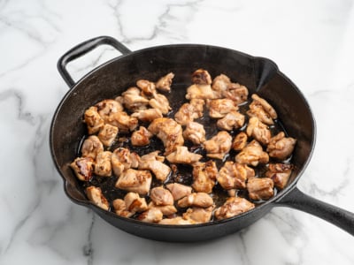 Chunks of browned chicken sizzle in a black cast iron skillet on a white marble countertop. Steam rises as the meat cooks evenly—perfect for a homemade Kung Pao Chicken recipe.