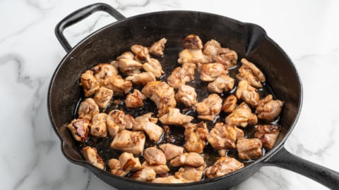 Chunks of browned chicken sizzle in a black cast iron skillet on a white marble countertop. Steam rises as the meat cooks evenly—perfect for a homemade Kung Pao Chicken recipe.