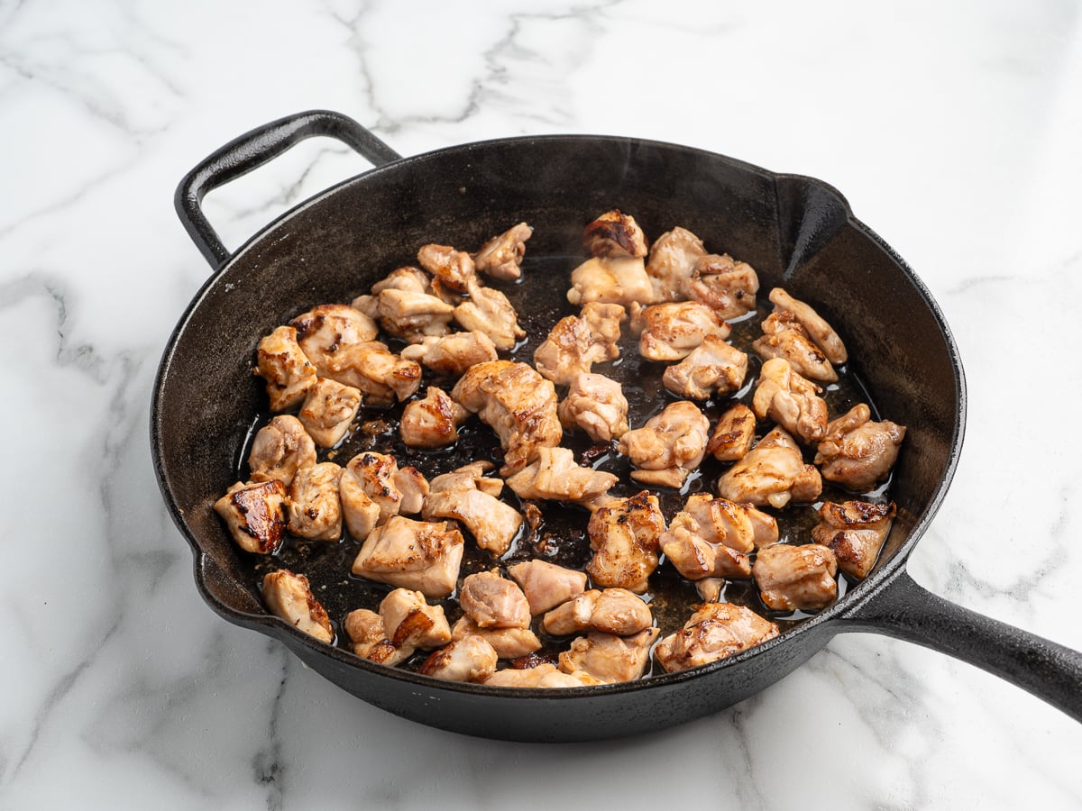 Chunks of browned chicken sizzle in a black cast iron skillet on a white marble countertop. Steam rises as the meat cooks evenly—perfect for a homemade Kung Pao Chicken recipe.