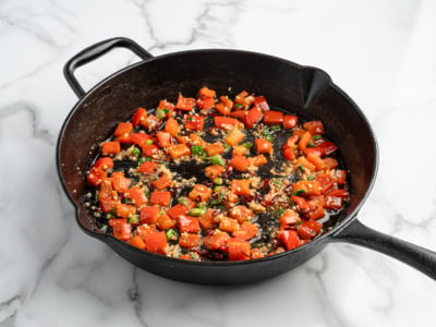 A black cast iron skillet on a marble surface contains sautéed diced red bell peppers, green herbs, and garlic. Reminiscent of a fresh Kung Pao Chicken recipe, the vegetables are lightly cooked and glistening with oil.