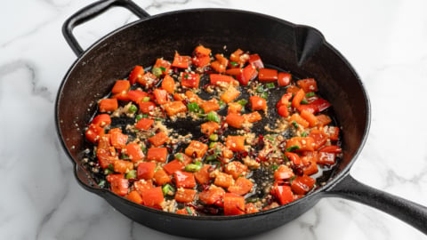 A black cast iron skillet on a marble surface contains sautéed diced red bell peppers, green herbs, and garlic. Reminiscent of a fresh Kung Pao Chicken recipe, the vegetables are lightly cooked and glistening with oil.
