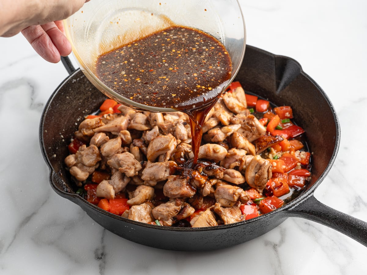 A hand pours a bowl of dark, seasoned sauce over cooked chicken pieces and chopped red bell peppers in a black cast iron skillet, creating a mouthwatering homemade kung pao chicken on a white marble surface.