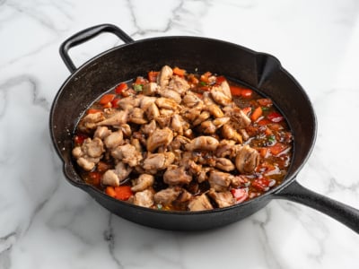 A black cast iron skillet on a marble surface contains cooked, seasoned chicken pieces mixed with chopped red bell peppers and a savory kung pao sauce, perfect for trying your own Kung Pao Chicken at home.