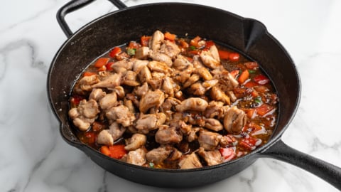 A black cast iron skillet on a marble surface contains cooked, seasoned chicken pieces mixed with chopped red bell peppers and a savory kung pao sauce, perfect for trying your own Kung Pao Chicken at home.