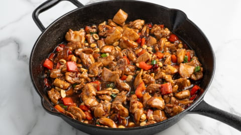 A cast iron skillet filled with cooked diced chicken, red bell peppers, peanuts, and green onions in a glossy brown sauce sits on a white marble surface—an irresistible look at the best kung pao chicken recipe.