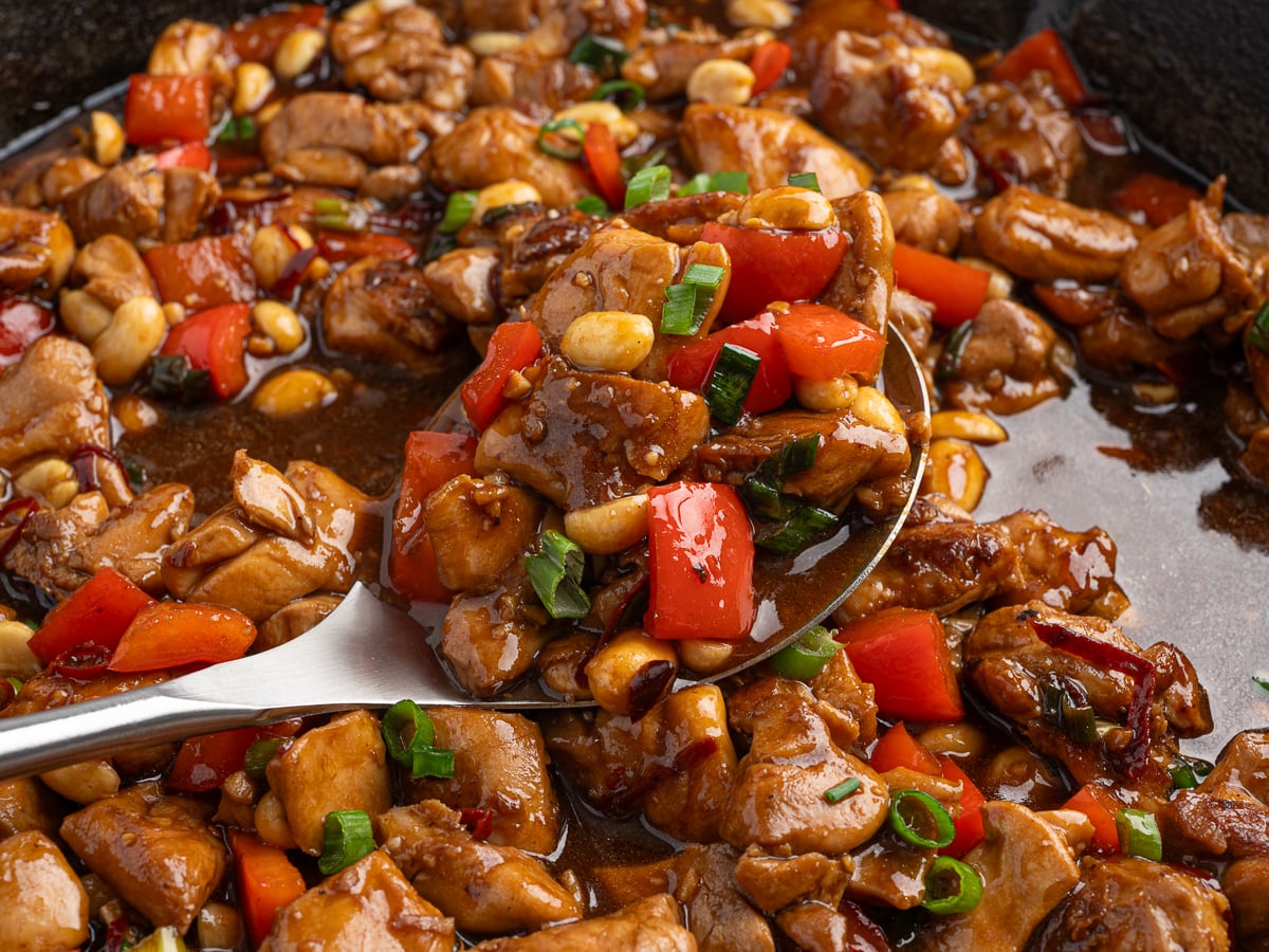 A close-up of a spoonful of homemade kung pao chicken with pieces of chicken, red bell peppers, peanuts, and green onions in a glossy brown sauce, surrounded by more kung pao in a pan.