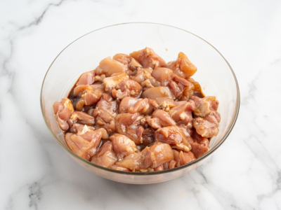 A glass bowl filled with marinated, diced raw chicken pieces sits on a white marble surface—perfect for starting the best kung pao chicken recipe.