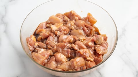 A glass bowl filled with marinated, diced raw chicken pieces sits on a white marble surface—perfect for starting the best kung pao chicken recipe.