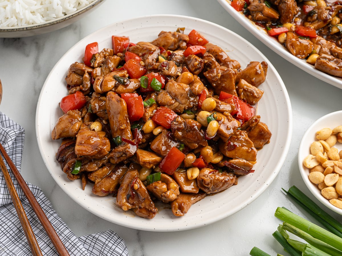 A plate of homemade Kung Pao Chicken featuring diced chicken, red bell peppers, peanuts, and green onions in a glossy sauce, served with rice and extra toppings on a marble surface.