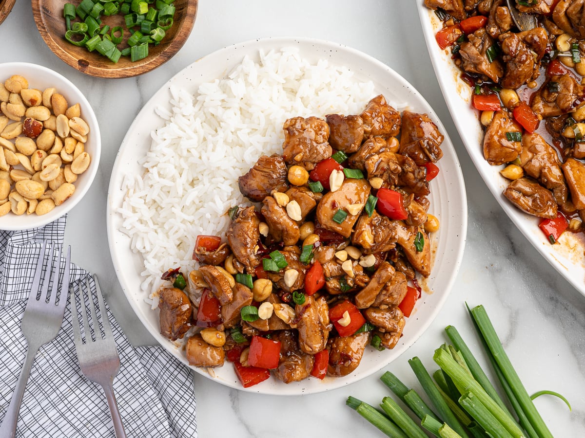 A plate of homemade kung pao chicken with white rice, red bell peppers, peanuts, and green onions, garnished with chopped green onions. Bowls of peanuts and green onions sit nearby, alongside two forks and a napkin.