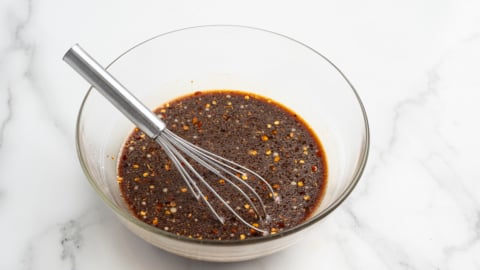 A glass bowl filled with a dark, speckled sauce or marinade, perfect for a homemade kung pao chicken recipe, sits beside a metal whisk on a white marble surface.