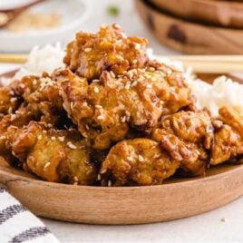 close up shot of a wooden plate of Sesame Chicken garnished with sesame seeds and served over white rice