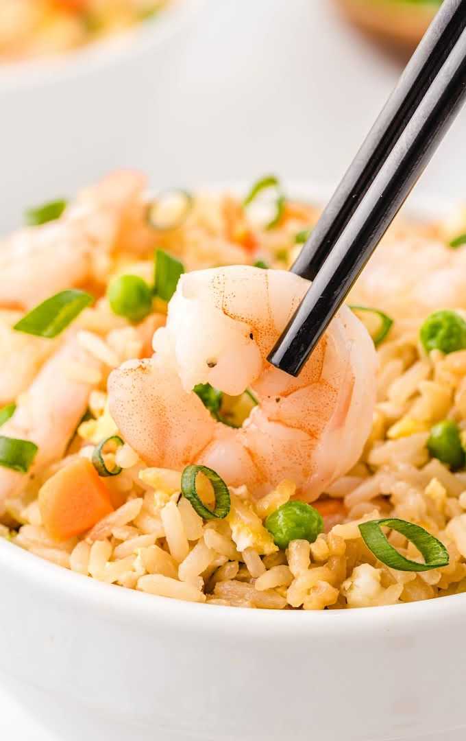 close up shot of shrimp fried rice garnished with green onions in a wooden bowl with shrimp being picked up with chop sticks