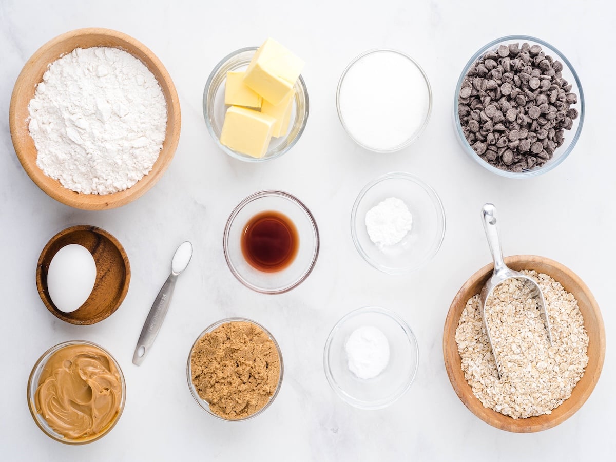Overhead view of baking ingredients in bowls for Peanut Butter Oatmeal Cookies: flour, butter, sugar, chocolate chips, oats, peanut butter, brown sugar, an egg, vanilla extract, salt, baking powder, and baking soda on a white surface.