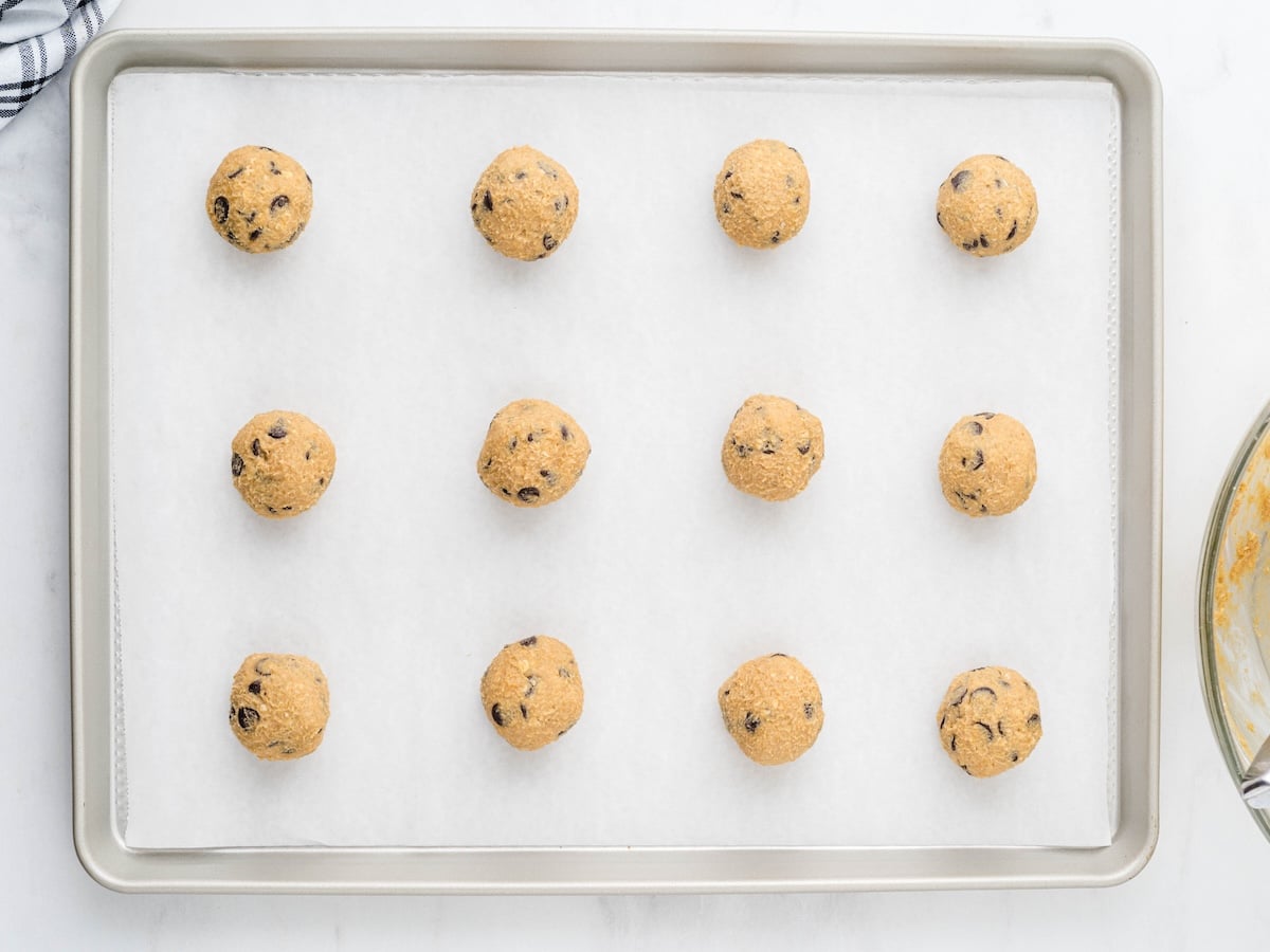 A baking sheet lined with parchment paper holds 15 evenly spaced balls of Peanut Butter Oatmeal Cookies dough, ready to be baked. The edge of a mixing bowl is visible on the right side.