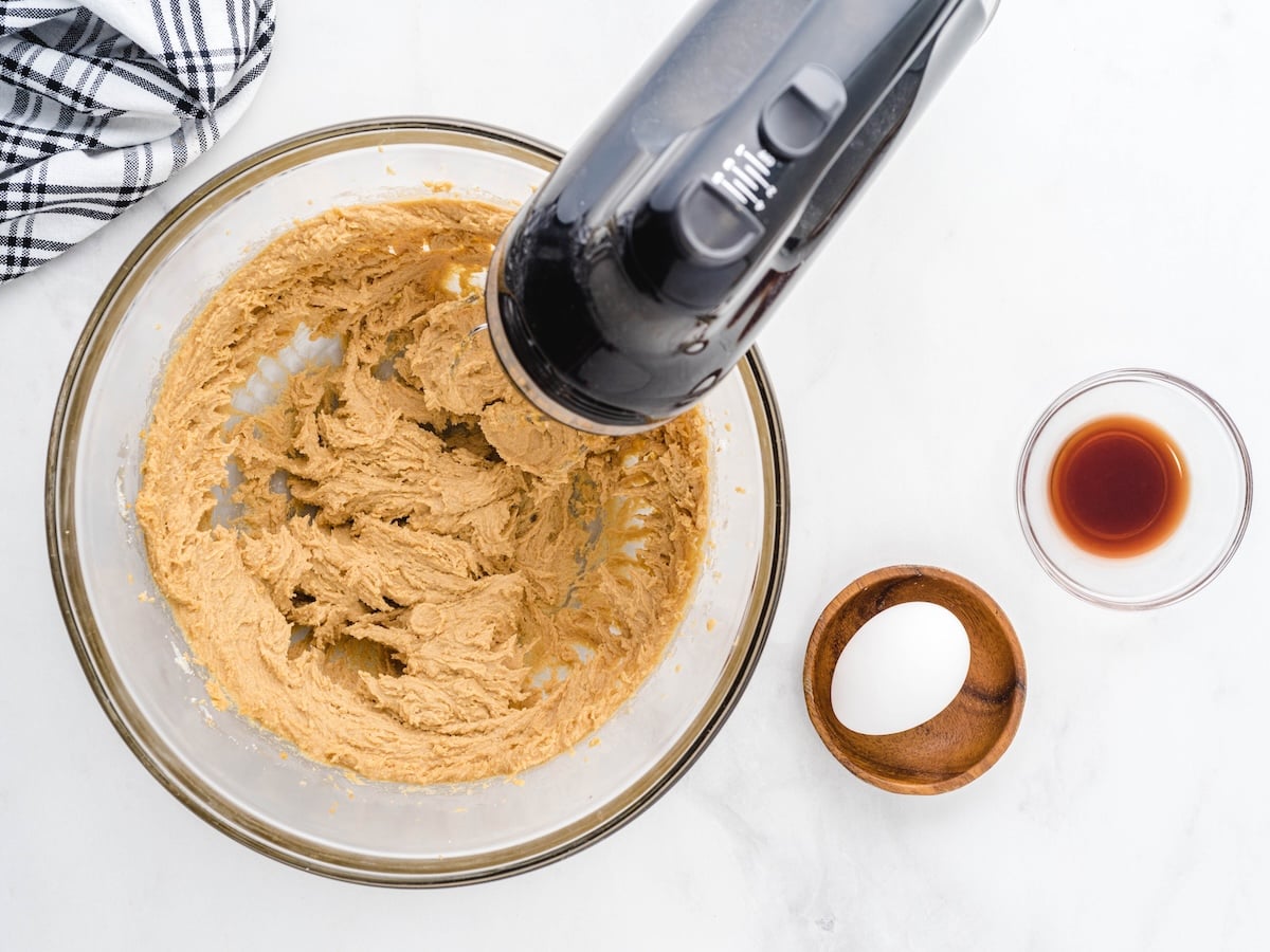 A glass bowl with cookie dough for Peanut Butter Oatmeal Cookies, an electric hand mixer inside, a whole egg in a wooden bowl, vanilla extract, and a black-and-white dish towel on a white surface.