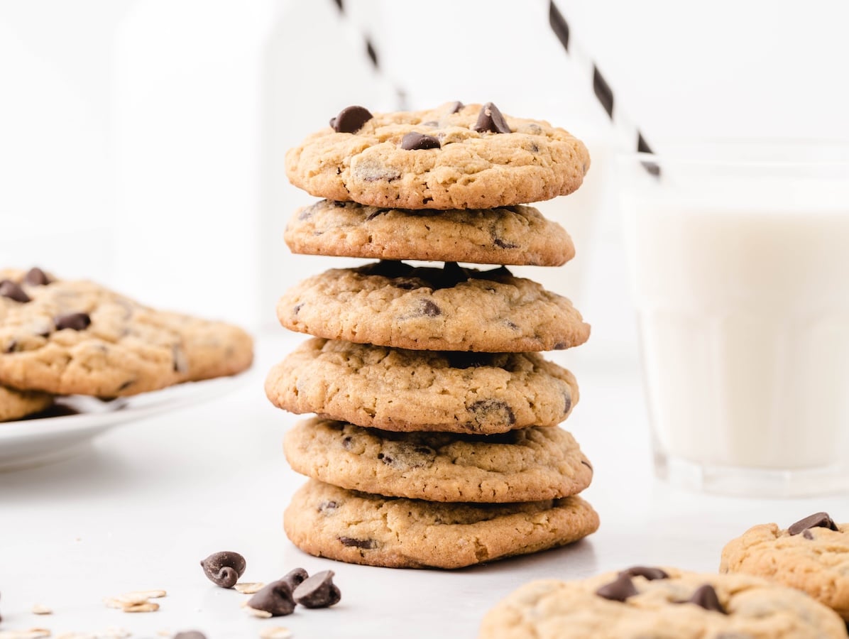 A stack of six Peanut Butter Oatmeal Cookies sits on a white surface, with a glass of milk, a plate of cookies, and a milk bottle with black striped straws in the background. Chocolate chips and oats are scattered nearby.