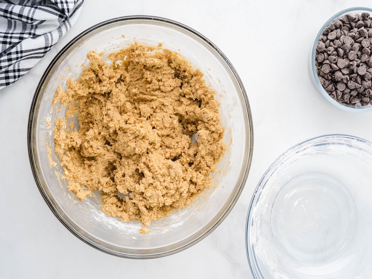 A glass bowl filled with cookie dough for Peanut Butter Oatmeal Cookies sits on a white surface. Nearby are an empty glass bowl, a small bowl of chocolate chips, and a black-and-white checkered cloth.