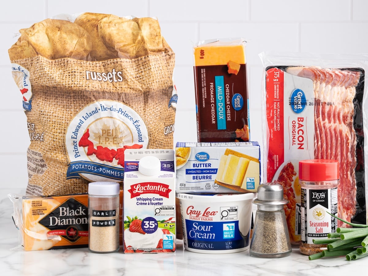 A variety of food ingredients on a counter, perfect for a Twice Baked Potato Casserole Recipe&mdash;russet potatoes, cheddar cheese, garlic powder, sour cream, whipping cream, butter, bacon, seasoning salt, black pepper, and green onions.