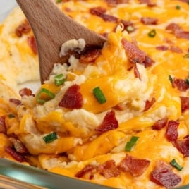 A wooden spoon scoops cheesy mashed potatoes from a glass baking dish—this Twice Baked Potato Casserole is topped with crispy bacon bits and chopped green onions for the perfect comfort food.