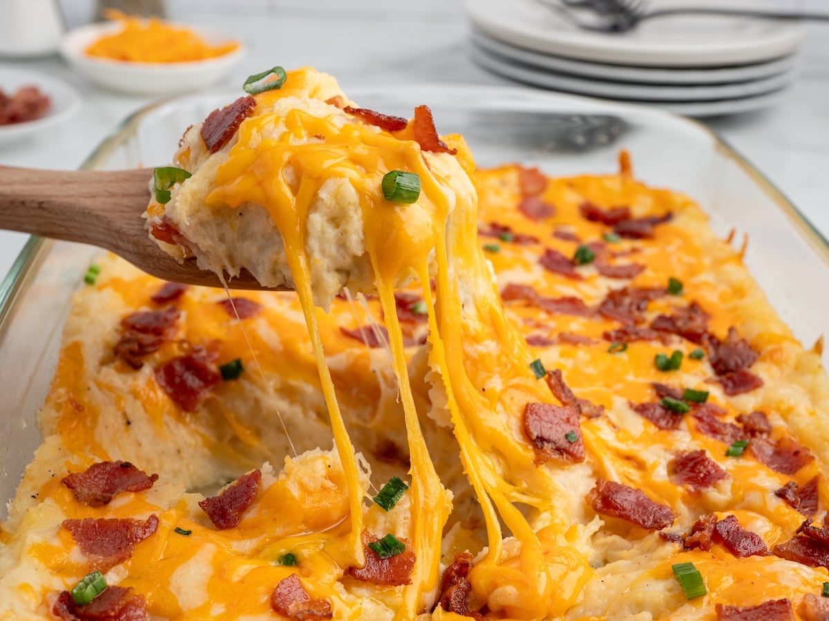 A wooden spoon lifts a serving of Twice Baked Potato Casserole topped with melted cheddar, crispy bacon, and green onions from a glass baking dish. Cheese stretches deliciously from this comforting Twice Baked Potato Casserole Recipe.
