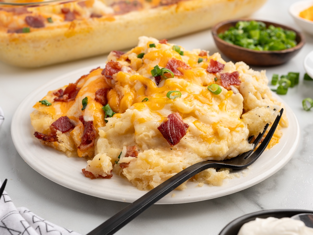 A plate of cheesy Twice Baked Potato Casserole topped with melted cheese, crispy bacon pieces, and chopped green onions, with a black fork and a casserole dish and condiments in the background.