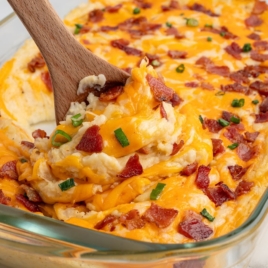 A wooden spoon scoops cheesy mashed potatoes from a Twice Baked Potato Casserole, topped with melted cheddar, crispy bacon pieces, and chopped green onions from a glass baking dish.