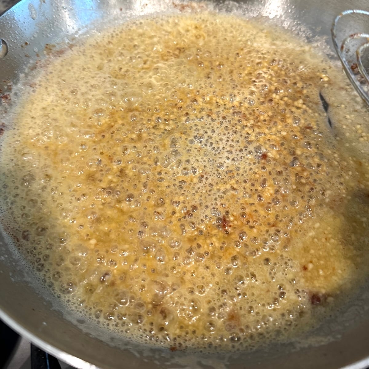 A close-up of a metal frying pan on a stove, filled with melted butter and bubbling golden-brown bits, shows the start of a delicious Creamy Chicken Pasta recipe being prepared.