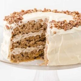 side shot of hummingbird cake with pecans on top with a slice being taken out of it on a cake dish