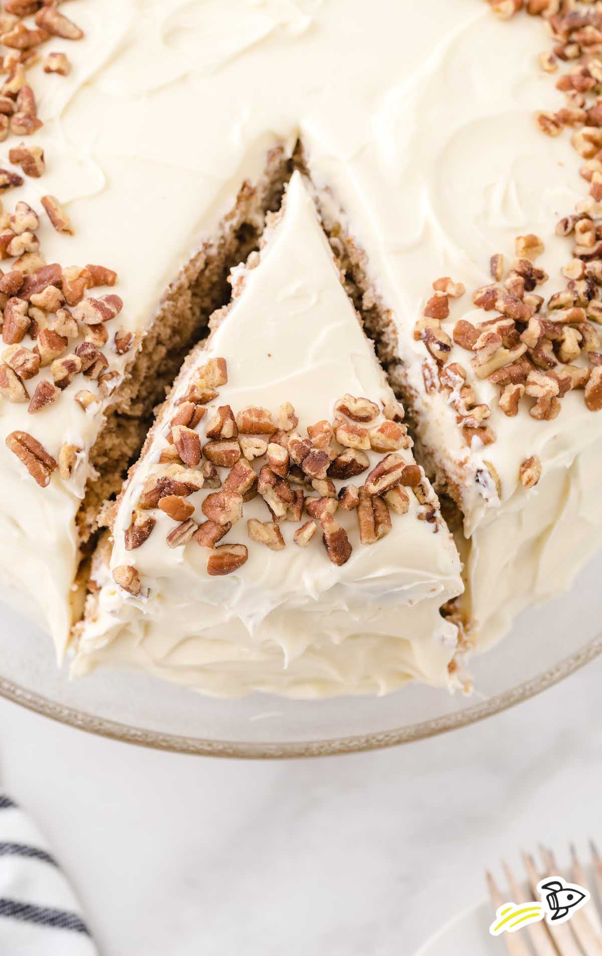 side shot of hummingbird cake with pecans on top with a slice cut out on a cake dish